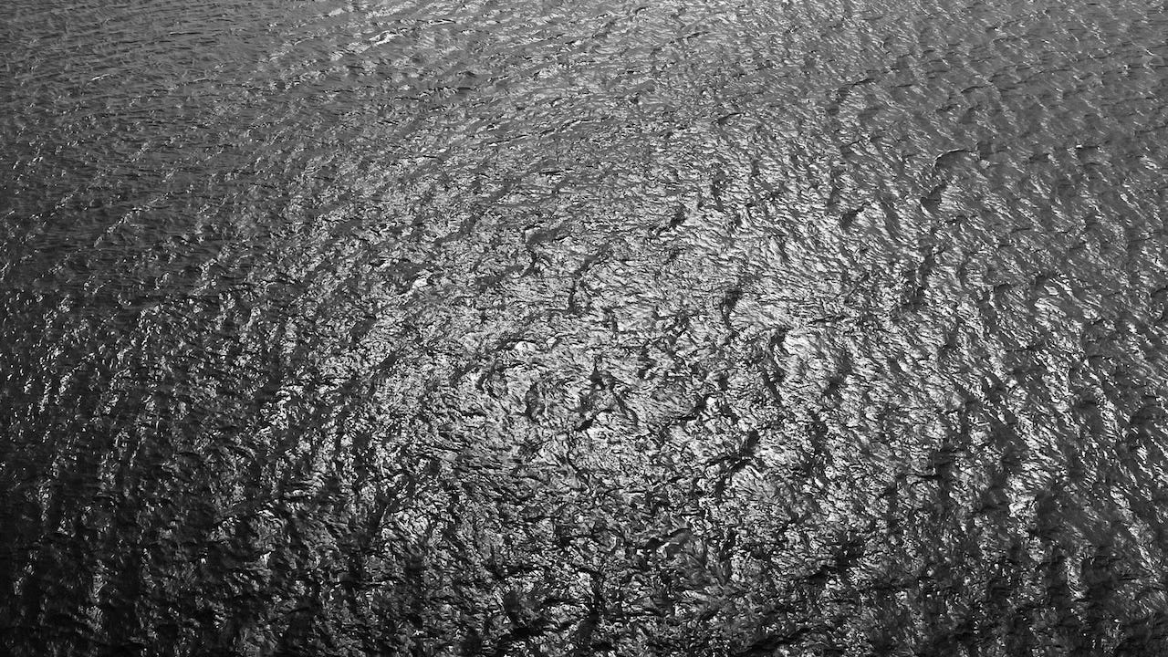 GPT-4o: A black-and-white aerial view of the ocean’s surface, showcasing intricate ripples and waves illuminated by light reflections. The textured water captures a sense of motion and natural beauty in this dynamic and timeless composition.