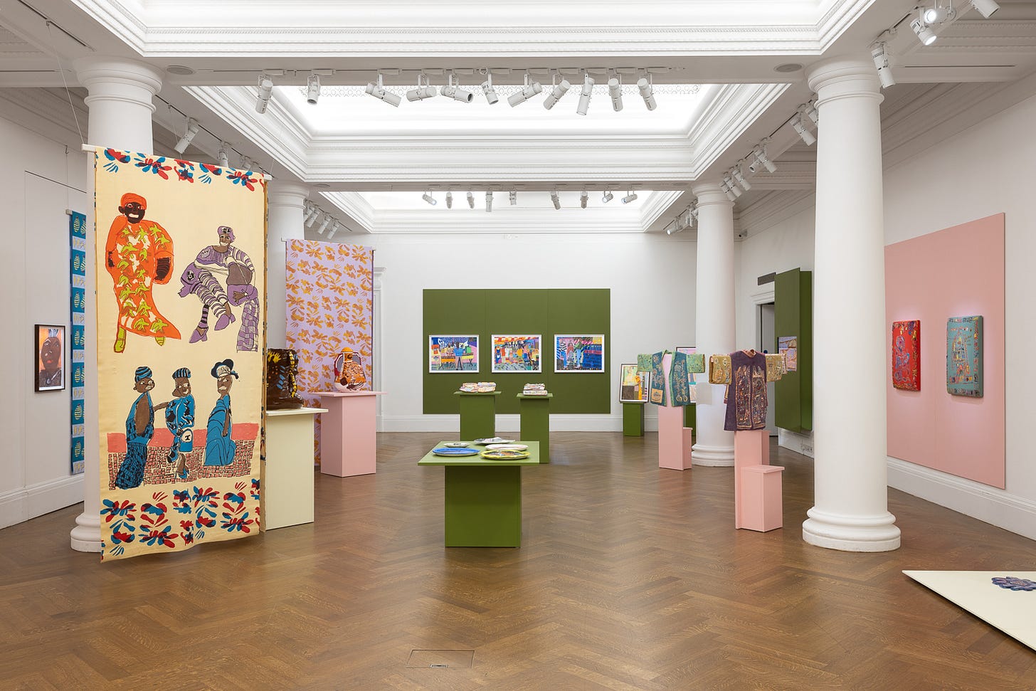 A photograph of a gallery space with plinths in the centre of the room displaying ceramics. Decorative fabric hangings and garments line the left and right. Around the room are framed artworks.