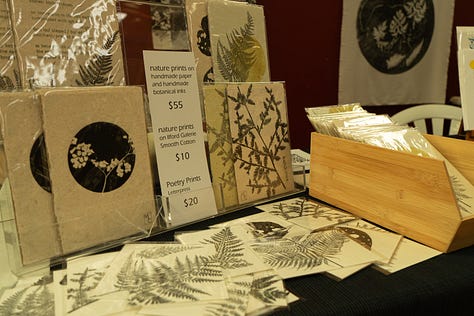 The table is covered in many different botanical and text based prints on different surfaces, including a fabric book. In the second photo I am standing at the stall, with two large fabric botanical print hangings on the wall behind me., In the first photo is a close up of the letterpress text test prints on calico. The text overlaps itself making a tall pillar of text overlapping and fading in and out of itself.
