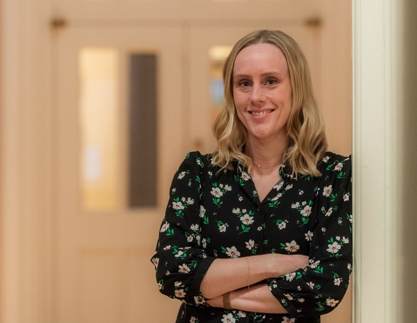Christina Lister, a white woman smiling with her arms folded, leaning against a wall in a corridor, wearing a floral dress.