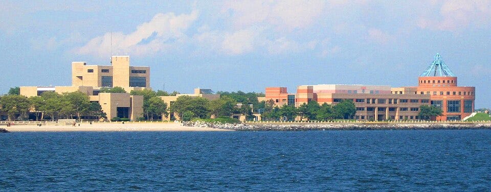 Kingsborough Community College’s brick waterfront campus seen across Jamaica Inlet, with a rocky shoreline, lawns, and a glass-fronted rotunda beneath a clear sky. Kingsborough Community College’s brick waterfront campus seen across Jamaica Inlet, with a rocky shoreline, lawns, and a glass-fronted rotunda beneath a clear sky.