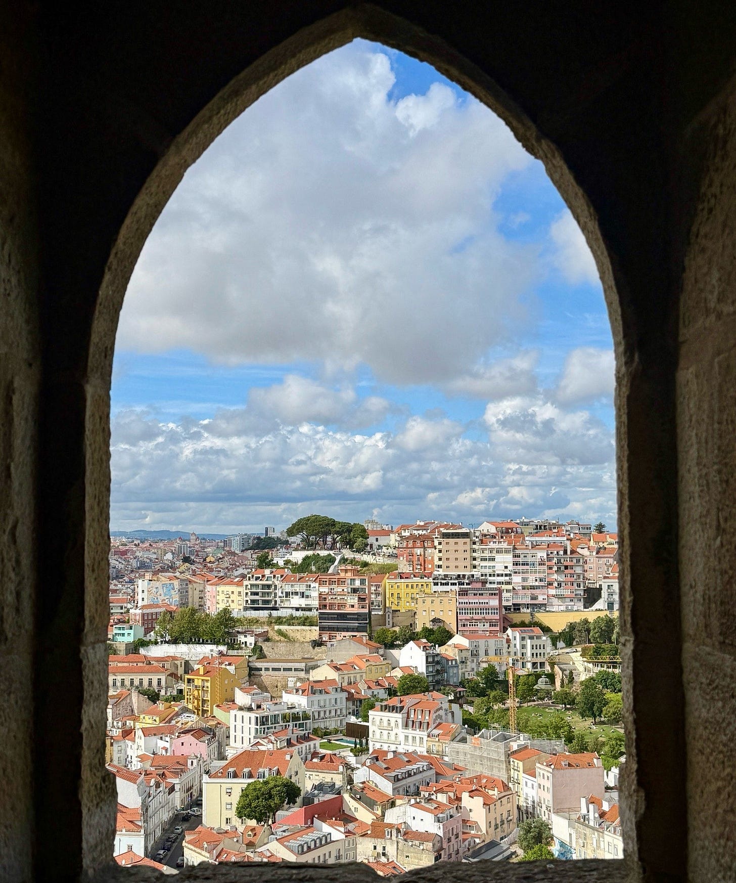 Lisbon's terracotta rooftops and pastel buildings seen through a pointed stone arch from an elevated viewpoint.