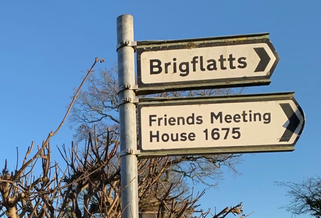 British road signs pointing to Briggflatts / Friends Meeting House 1675 against a blue winter sky, hedge and tree