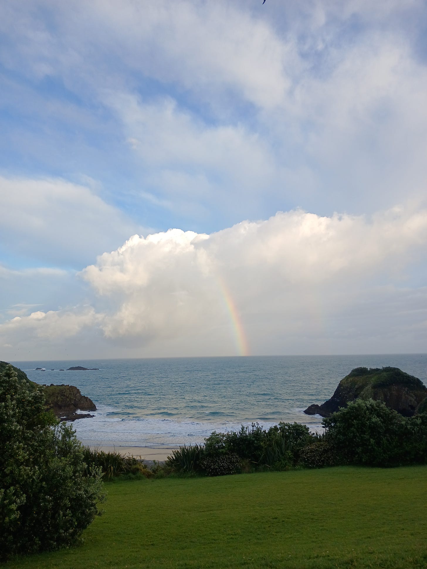 Image of the sea from a grassy clifftop. the sky is cloudy and there is one clear rainbow, and a second much fainter one