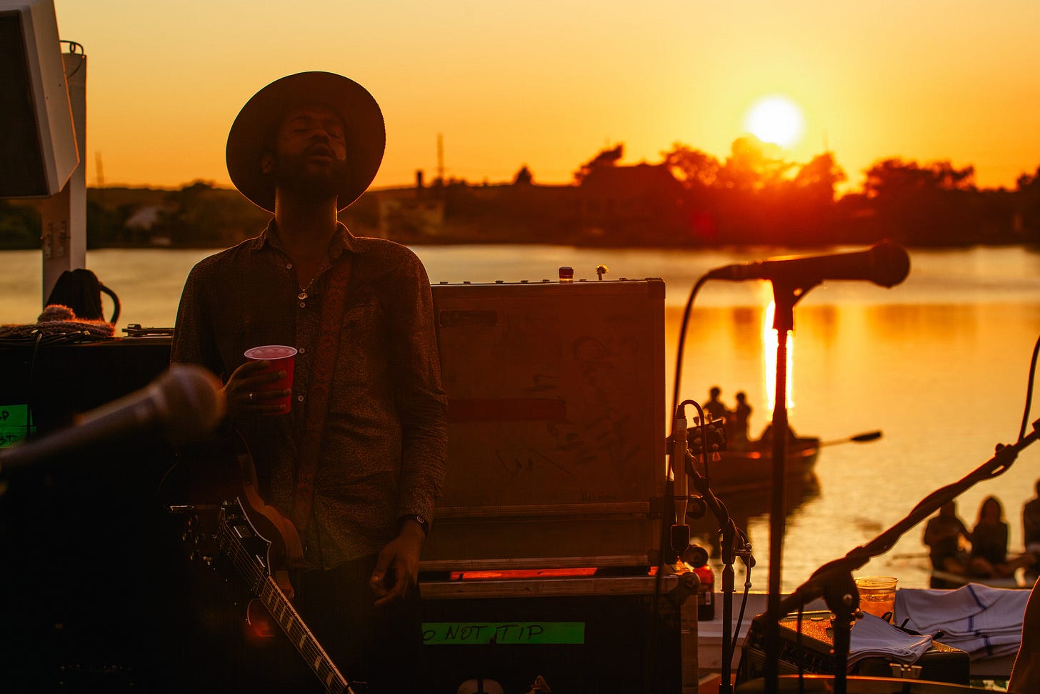 Gary Clark Jr. stands on an outdoor stage at sunset, holding a red cup with his eyes closed. The setting sun creates a golden glow over the water behind him, and the silhouettes of people in a boat are visible.