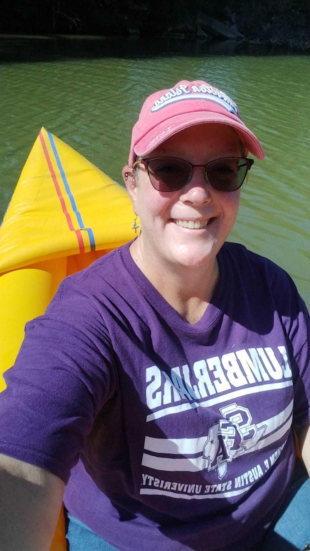 A selfie photo of a middle-aged woman wearing sunglasses, a pink ball cap and purple t-shirt. She is floating on a creek in a kayak.