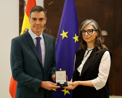 Sanchez and Albanese hold an award box together in front of Spanish and EU flags