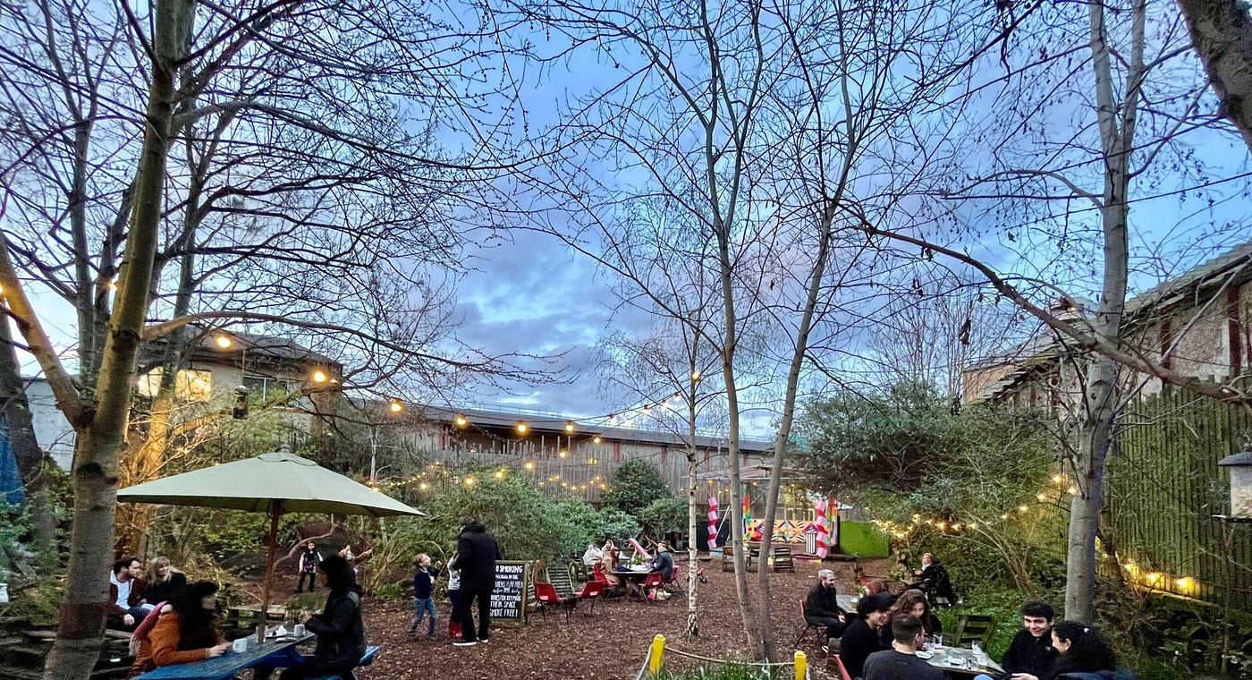 A natural city garden with trees, fairy lights and people sitting at tables A natural city garden with trees, fairy lights and people sitting at tables