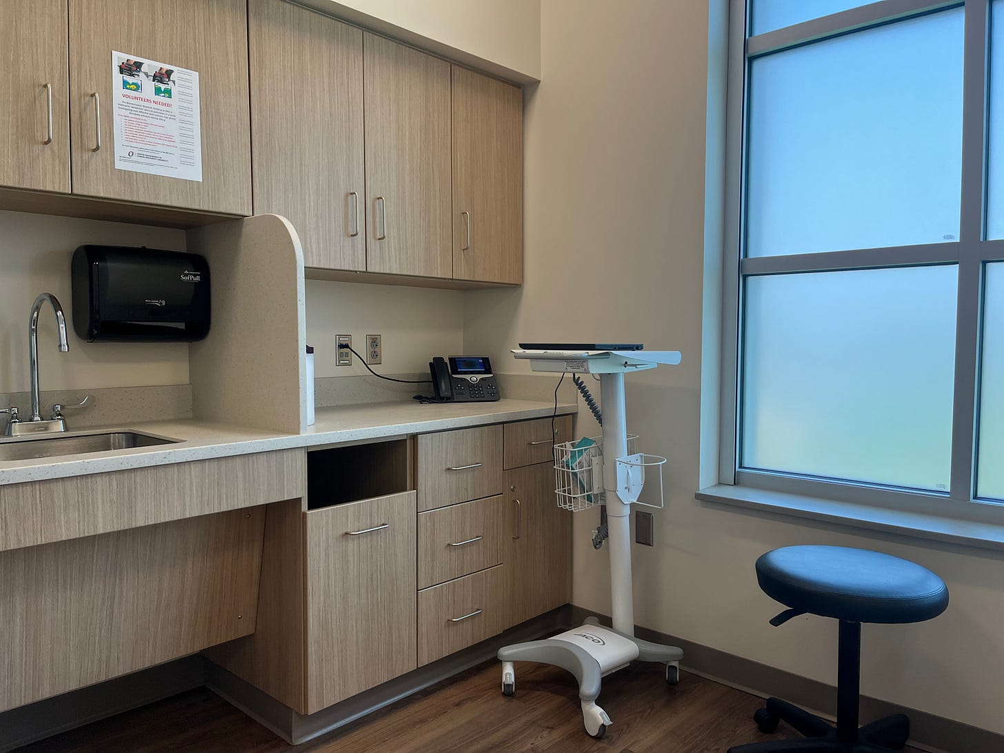 Doctors office with clinical cabinets, sink and fountain, laptop stand, and stool.