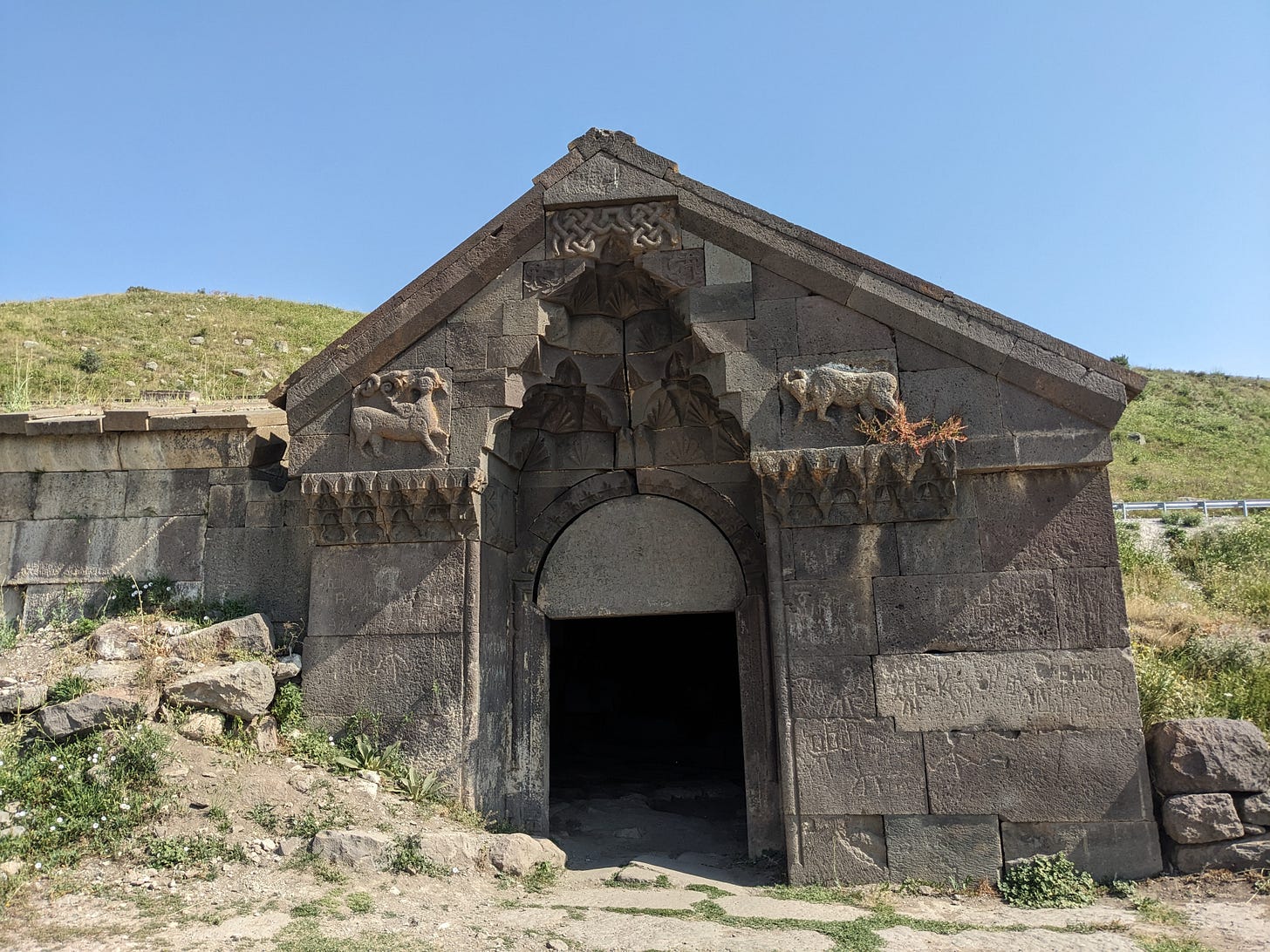 A grey stone building has an arched doorway with animals carved on each side. It is nestled in a meadow on a green hillside with blue sky behind.  