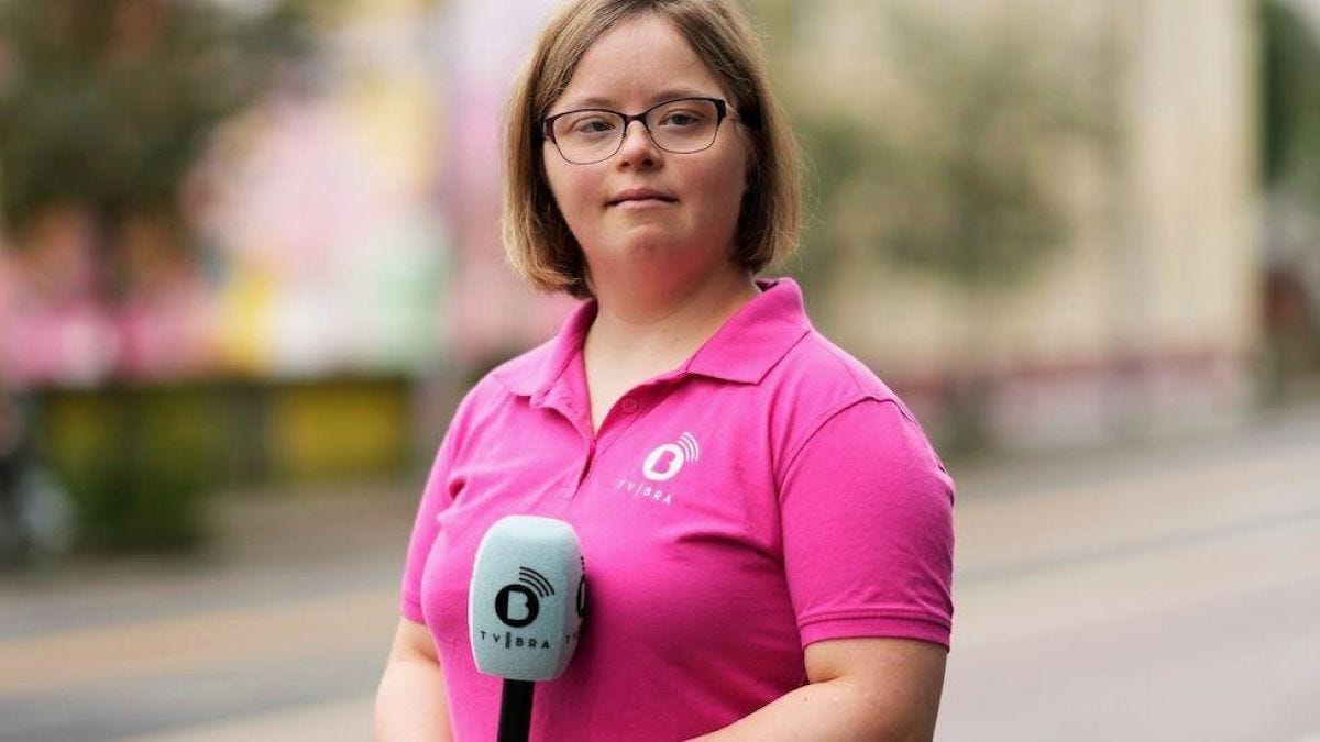 A white woman with Down Syndrome holds a broadcast microphone outside on a street. She wears a pink polo shirt and glasses.