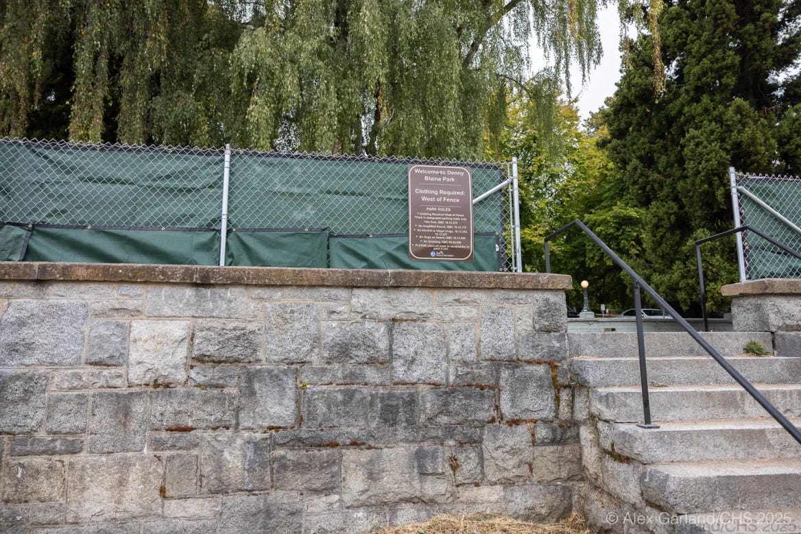 A stone stairway leads up to a new chain-link fence draped with green tarps at Denny Blaine Park, with a posted sign dividing clothing-required and clothing-optional areas.