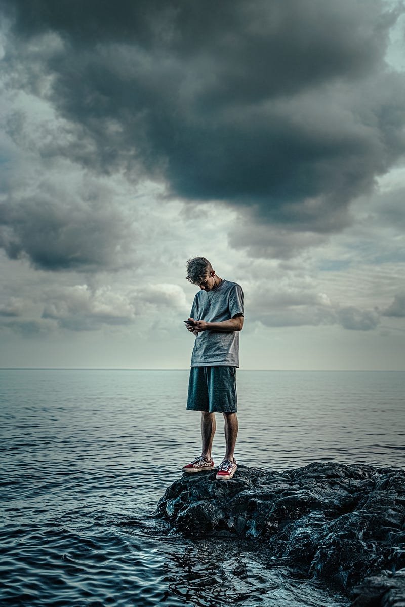 An escapist at the edge of a rock near an ocean turning to a phone. An escapist at the edge of a rock near an ocean turning to a phone.