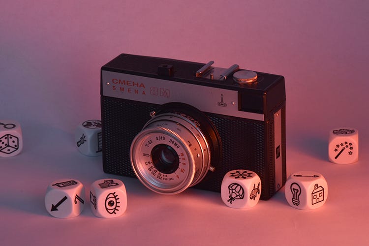 Boxy point-and-shoot camera surrounded by six-sided dice with icons of random objects, like an eye, arrow, cell phone, hot air balloon, fish, light bulb, wand, house, magnifying glass, and die.