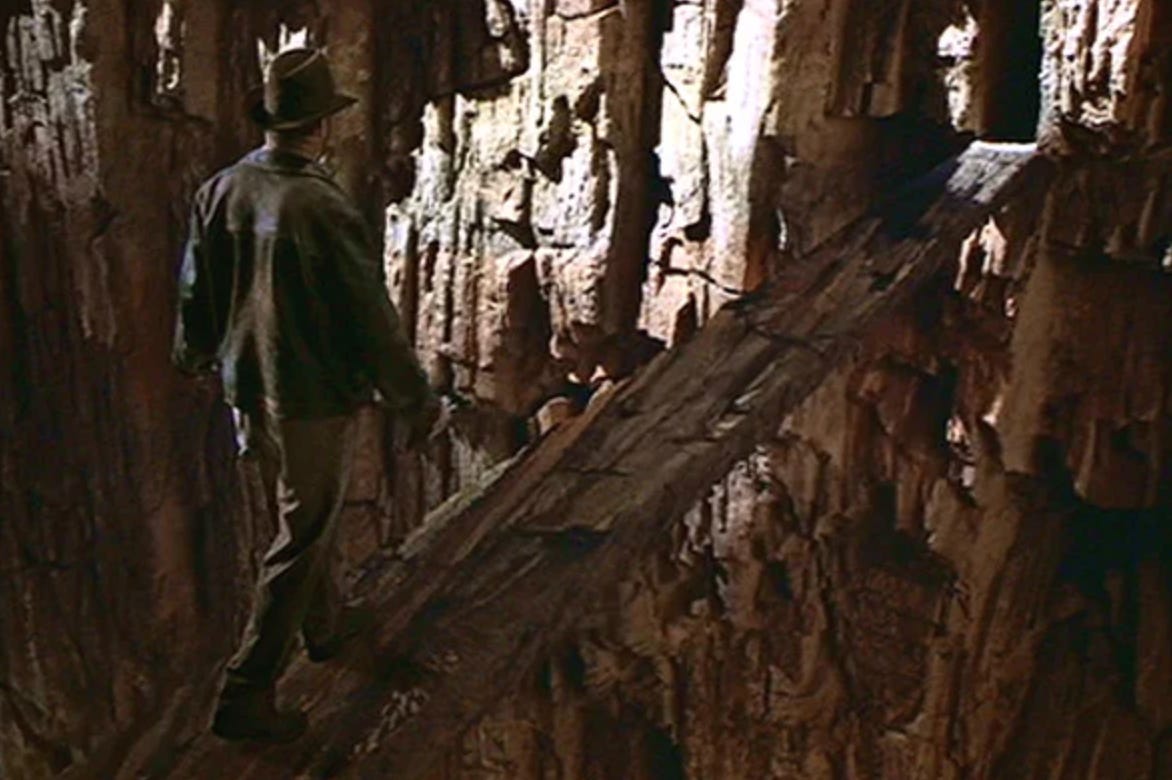 Indiana Jones in his brown leather jacket and fedora stands at the edge of a stone temple entrance, looking down at a chasm with tan and beige rock walls that camouflage the invisible bridge below Indiana Jones in his brown leather jacket and fedora stands at the edge of a stone temple entrance, looking down at a chasm with tan and beige rock walls that camouflage the invisible bridge below