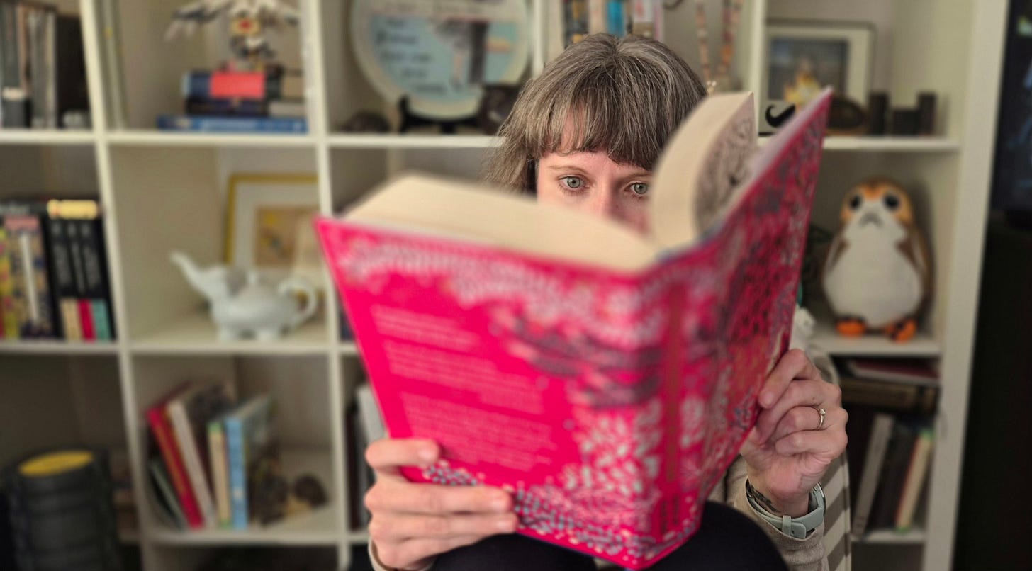 white woman reading large book with pink cover in front of bookcase white woman reading large book with pink cover in front of bookcase
