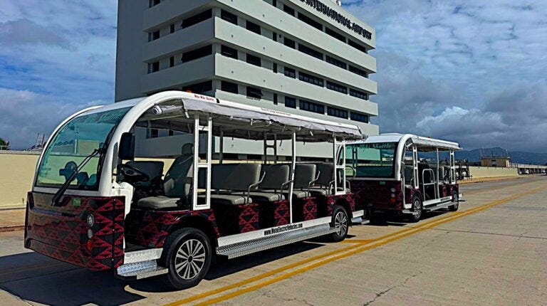 An electric tram parked on the tarmac at Honolou airport. The sun is shining and there is a mountain peak in the distance.