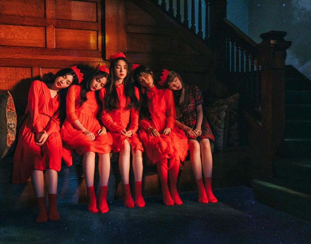 Red Velvet members Joy, Yeri, Irene, Seulgi, and Wendy dressed in red sitting together on a bench.