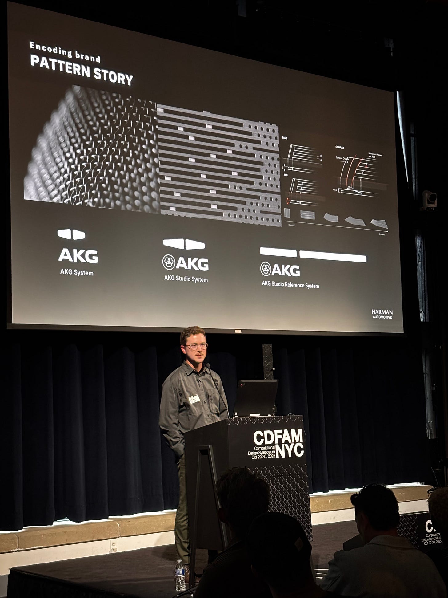 A speaker presenting at the CDFAM NYC 2025 event, with a screen displaying the AKG branding and system designs in the background.