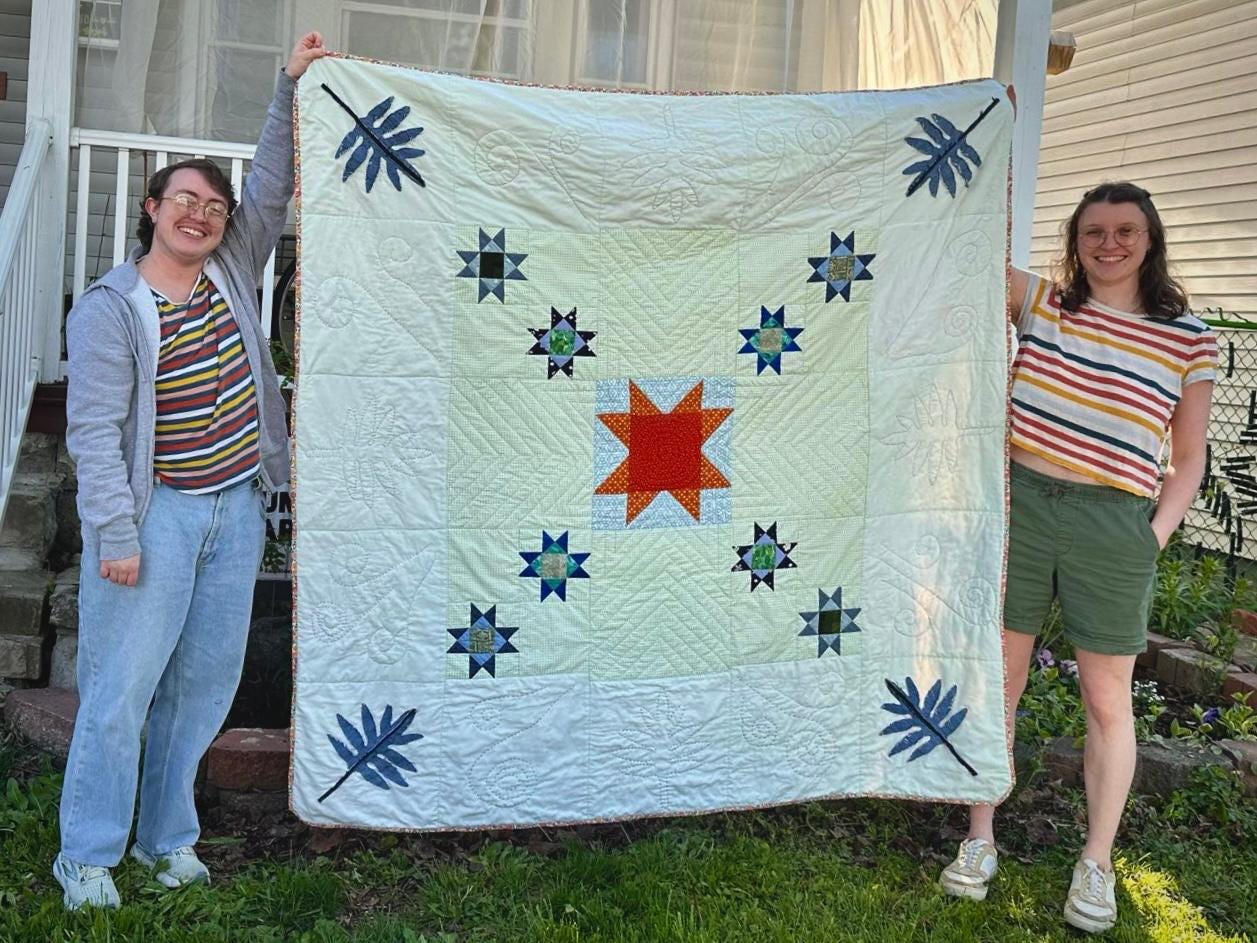 Image Description: Emory and Jo hold up a quilt they worked on with two other local quilters.
