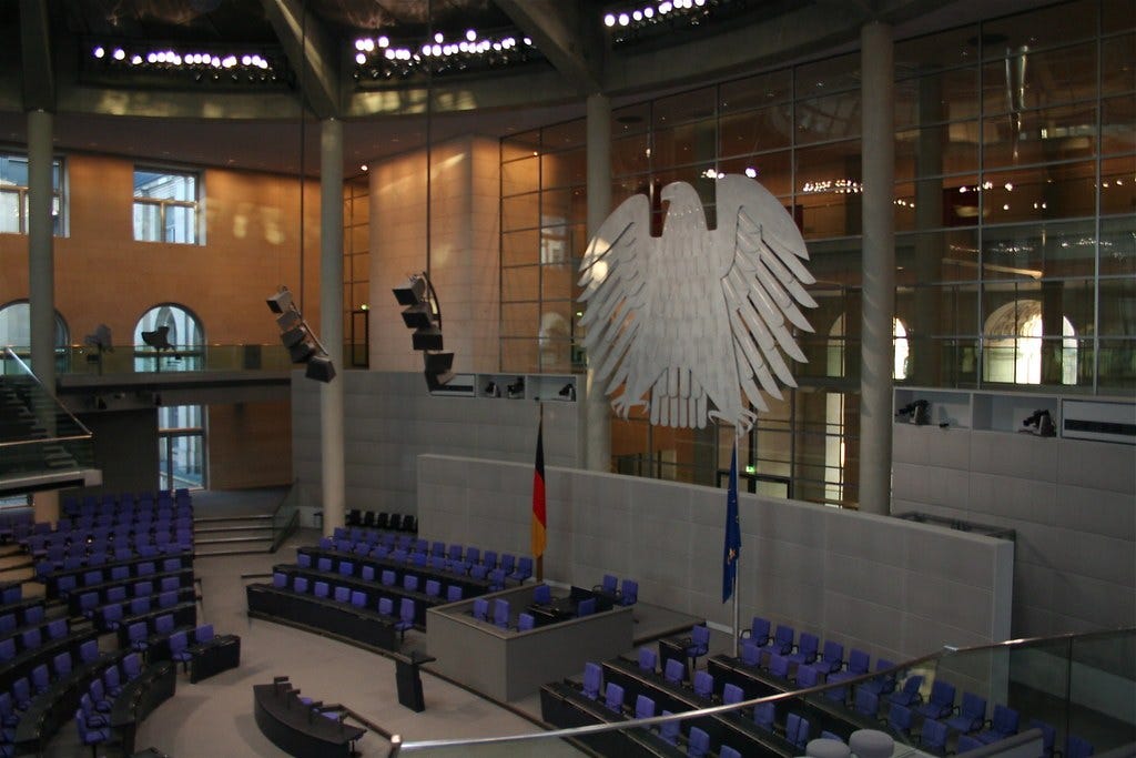 Rows of blue seats face a lectern beneath a large silver eagle in a glass-walled parliamentary chamber. Rows of blue seats face a lectern beneath a large silver eagle in a glass-walled parliamentary chamber.