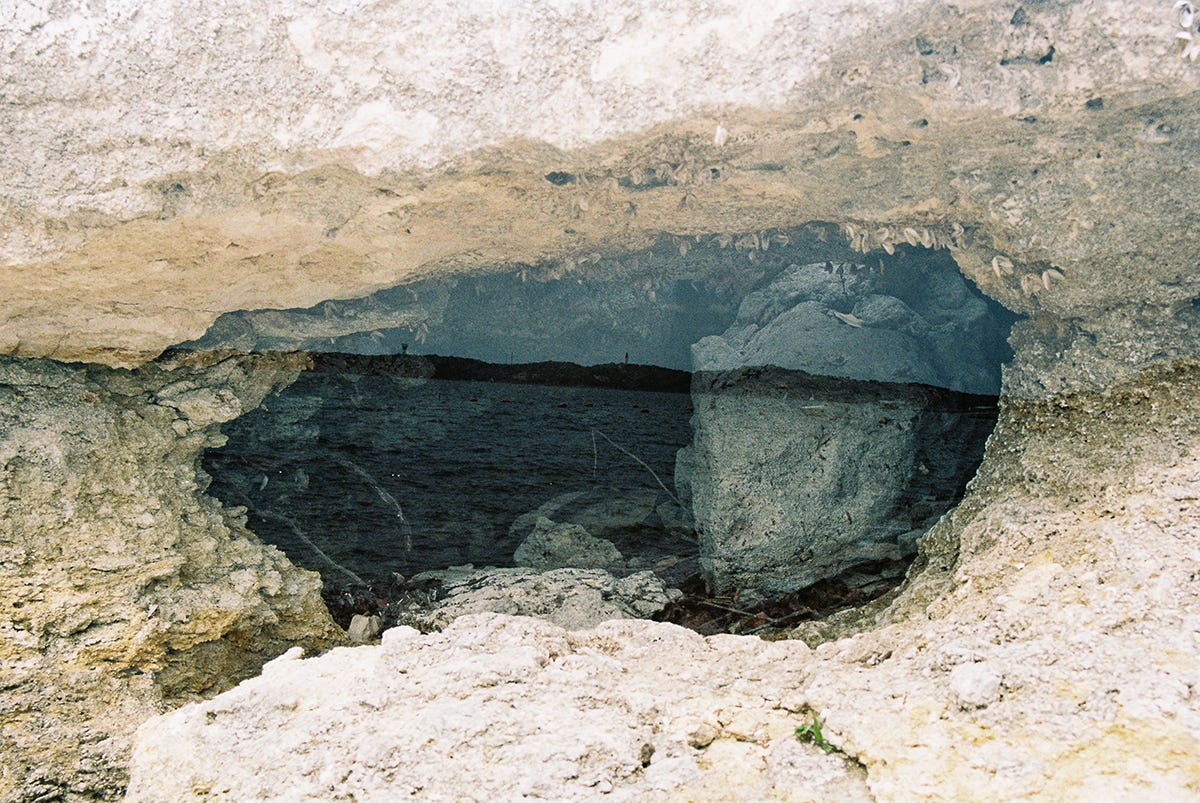 A double exposure of water’s horizon inside the circular mouth of a cave. A stripe of dark cuts through the mouth like an iris, gives the whorl the look of an eye. A stone in the opening of the cave situates itself like a pupil. The whole watches, looks right, and the crease of clams fringing the top become lashes, the column of sand becomes the bridge of a nose. The bright curve a bone, and suddenly, a cheek appears.