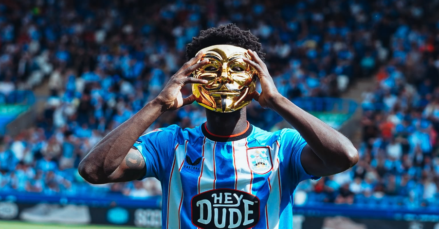 A Huddersfield Town player holds a golden mask to his face, while the crowd looks on in the background. A Huddersfield Town player holds a golden mask to his face, while the crowd looks on in the background.
