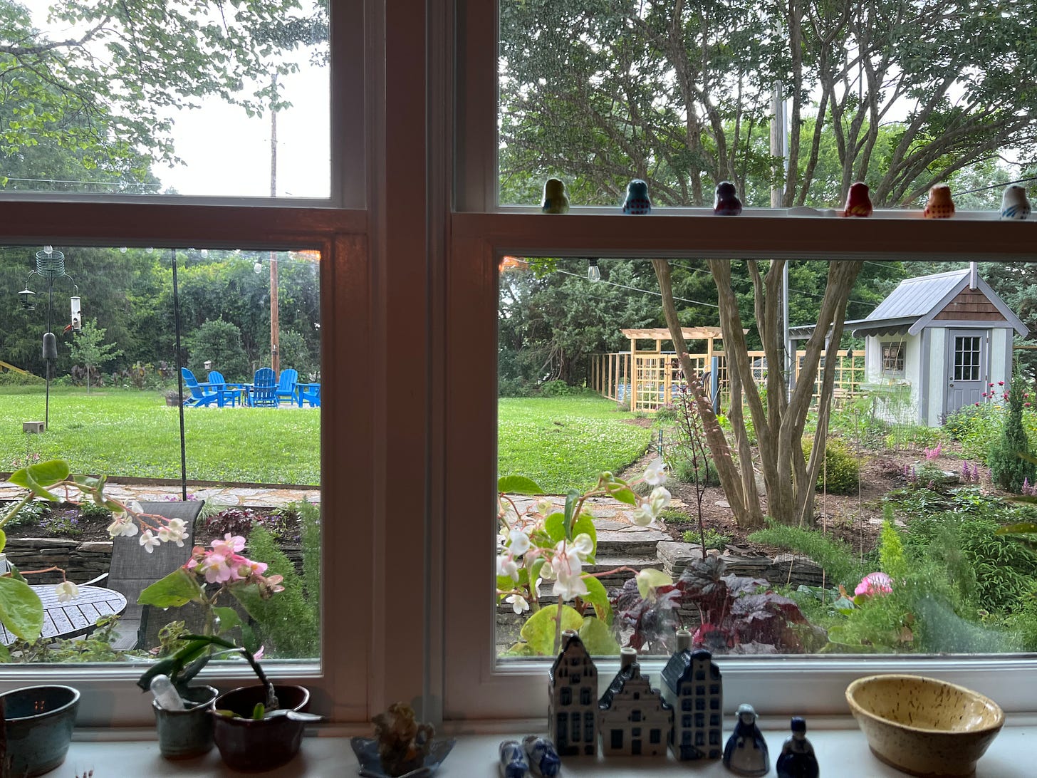 looking out a window at a green backyard with some blue chairs around a firepit and a fenced garden on the right side
