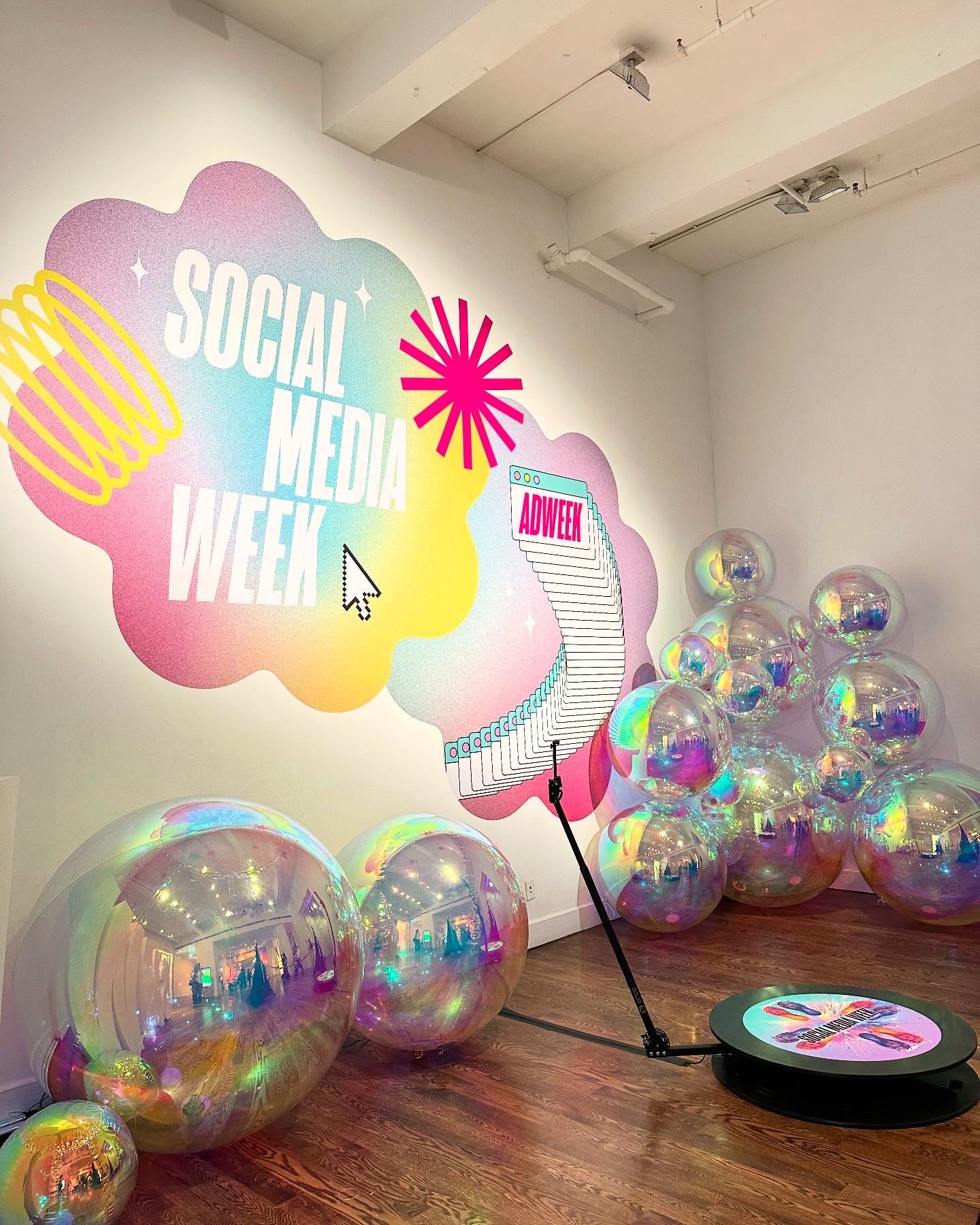 Wall sign that reads: Social Media Week. In the foreground is a photo station with silver balloons