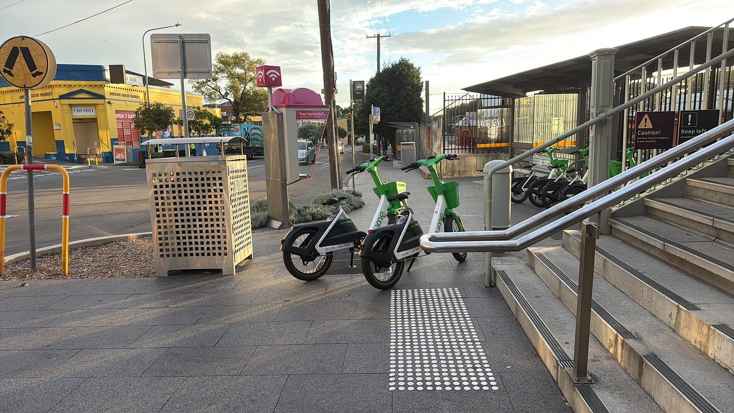 Two Lime bicycles block the footpath at entrance to a train station. Two Lime bicycles block the footpath at entrance to a train station.
