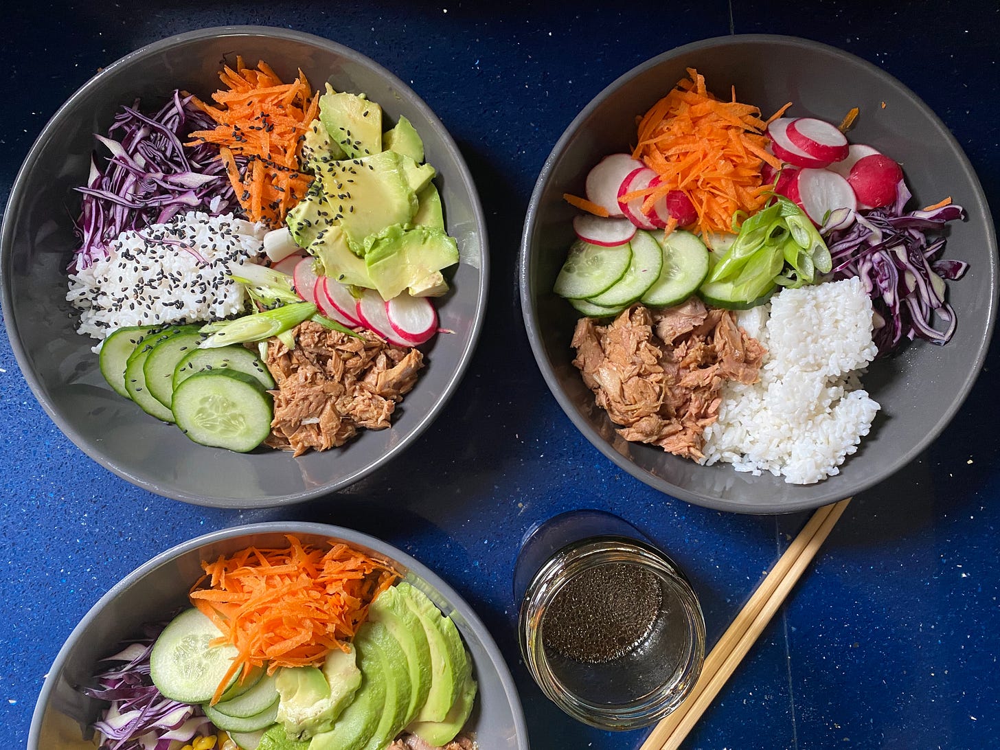 Three grey bowls on a blue quartz worktop. Each bowl is filled with chopped and shredded vegetables, some rice and tuna. A jar of sauce is next to them, and a set of chopsticks.