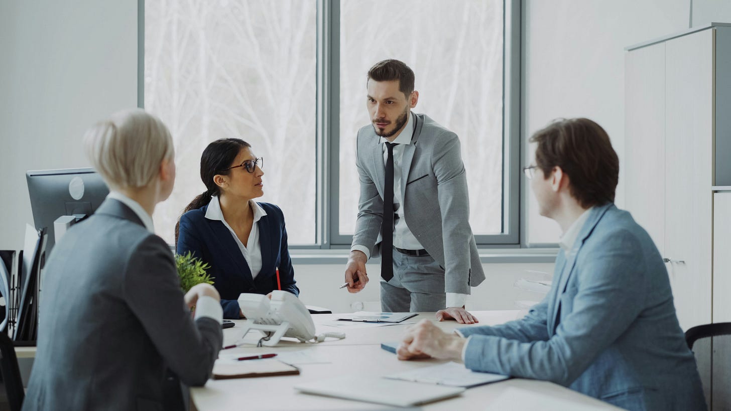 A person in a grey suit is standing by a desk, talking to three colleagues. They are all in suits. Two women and one man. It looks like they are discussing work and the leader is updating them