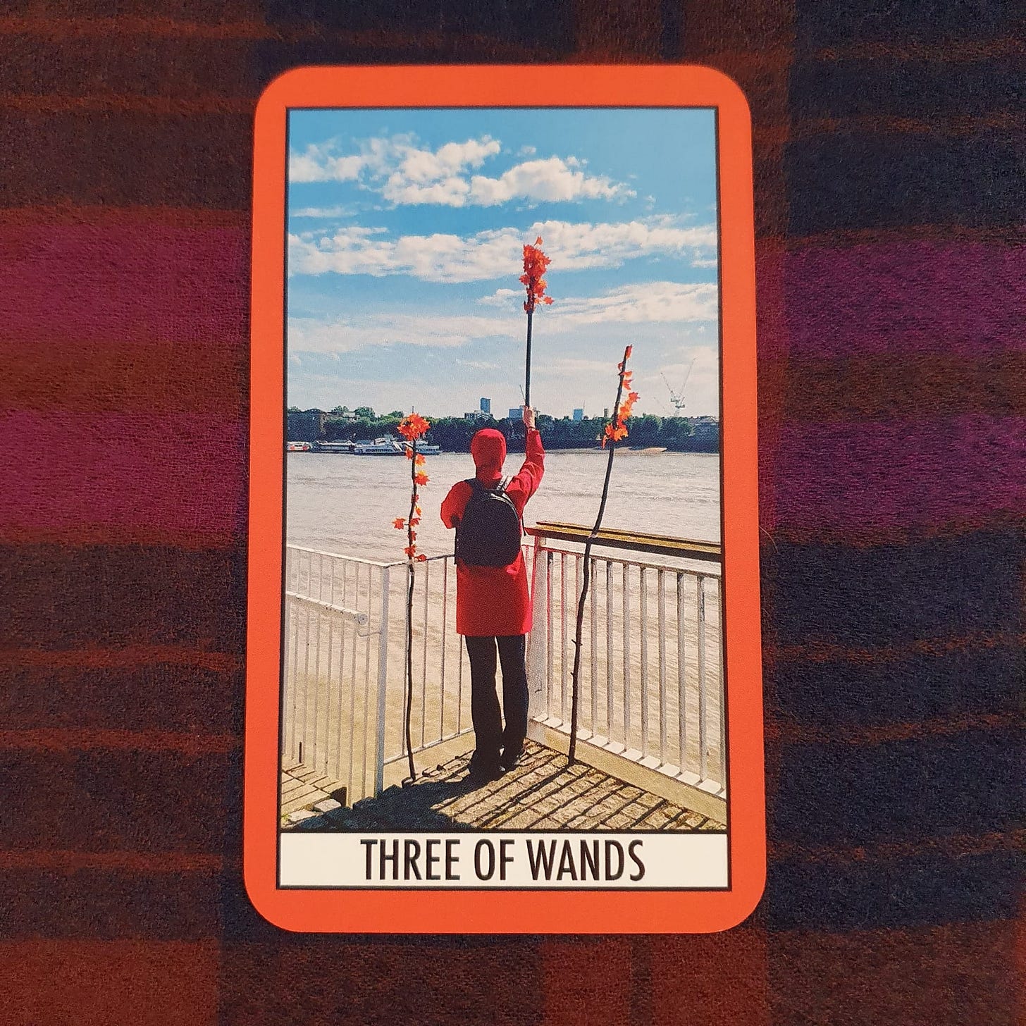 Three of Wands card from The Theatre Deli Tarot, showing a figure in red facing a river or open water with three decorated staffs raised against a bright blue sky. Tarot card pulled during Amy Twigg’s Tarot DMs interview to explore expansion, creative momentum, future books and hopeful next steps. Three of Wands card from The Theatre Deli Tarot, showing a figure in red facing a river or open water with three decorated staffs raised against a bright blue sky. Tarot card pulled during Amy Twigg’s Tarot DMs interview to explore expansion, creative momentum, future books and hopeful next steps.