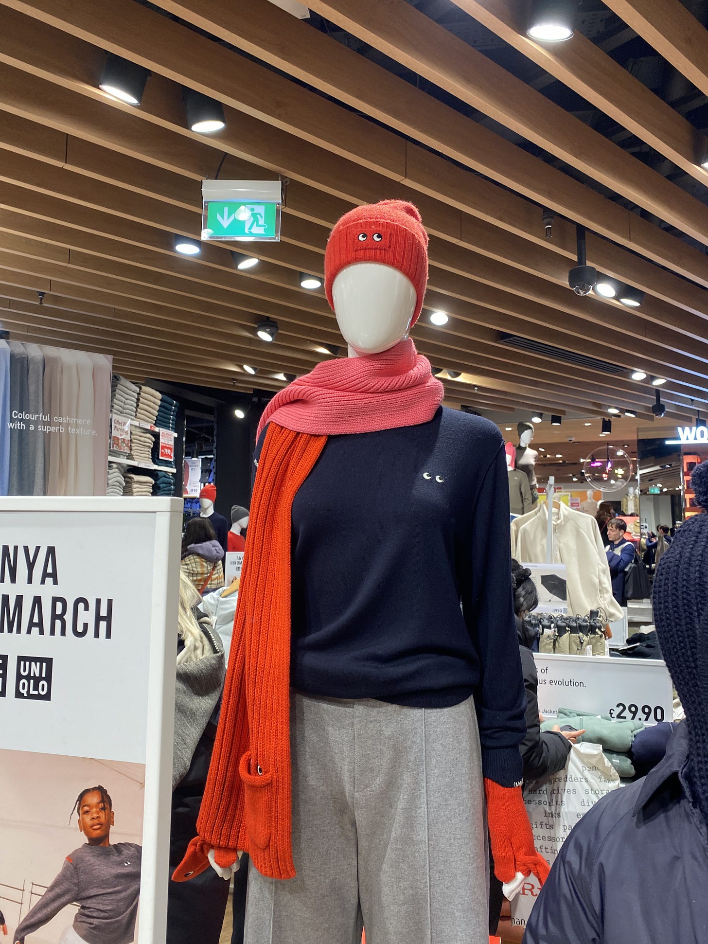 A Uniqlo manequin wearing Anya Hindmarch X Uniqlo which consists of a pink and red scarf with googly eyes, navy sweater with googly eyes, red beanie with googly eyes, red gloves and grey joggers