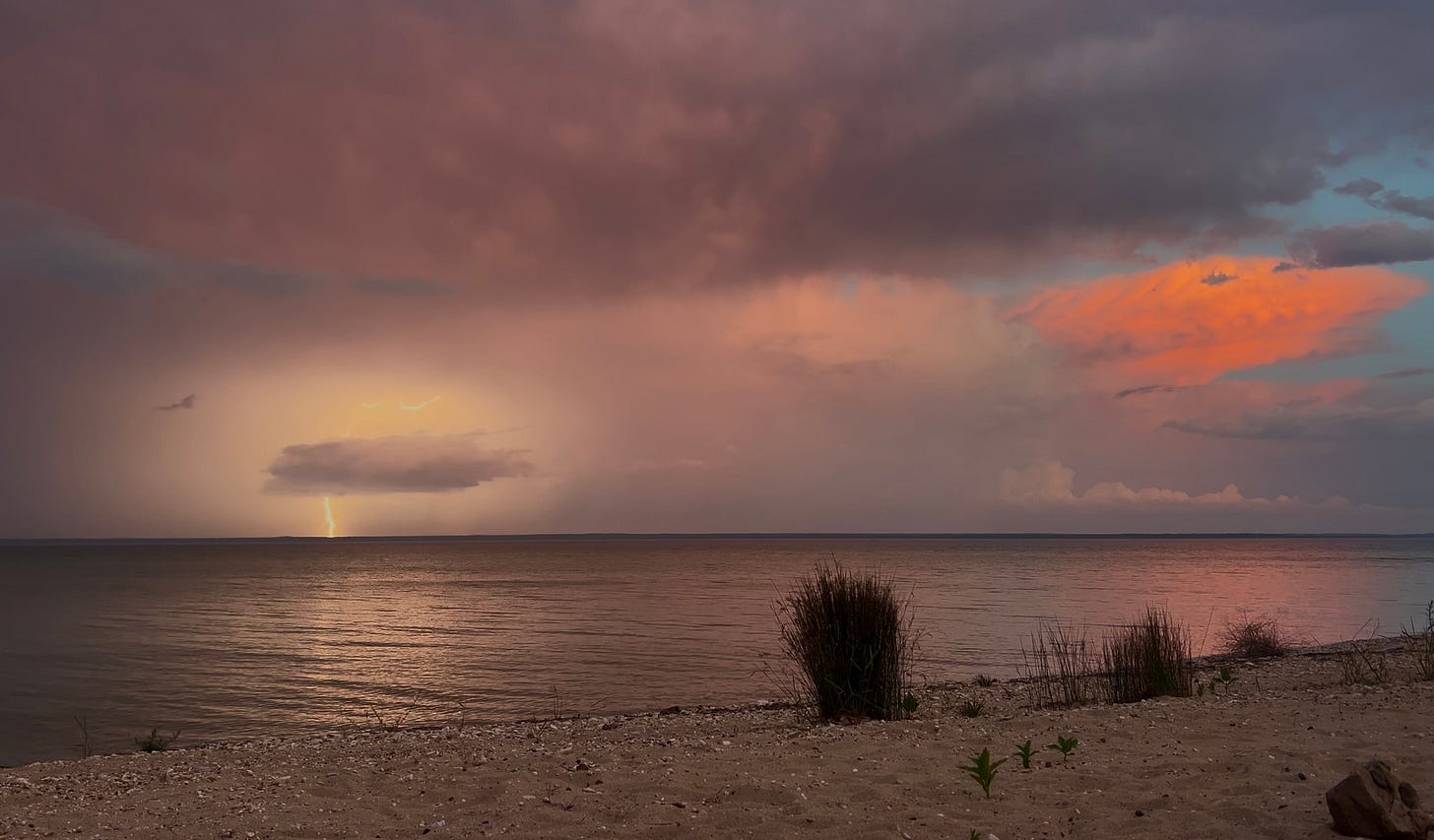 a stormy scene over a flat lake. a big pink shelf cloud on the left, and a bright white flash of lightning goes to the left and down to the water behind a small gray cloud on the left side of the image.