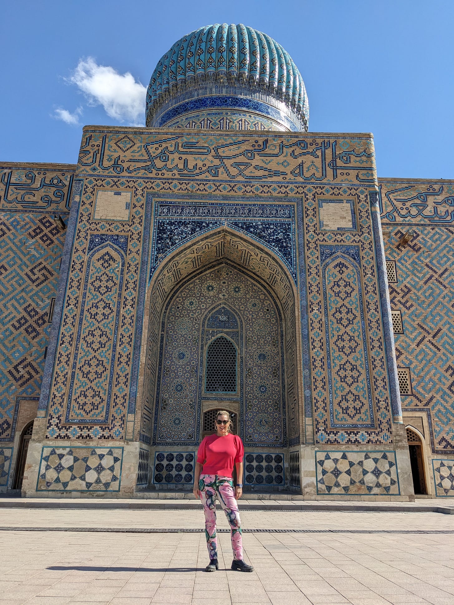 White woman wearing sunglasses, a pink sweater and tie-dyed jeans standing in front of a beautifully tiled Central Asian mosque.