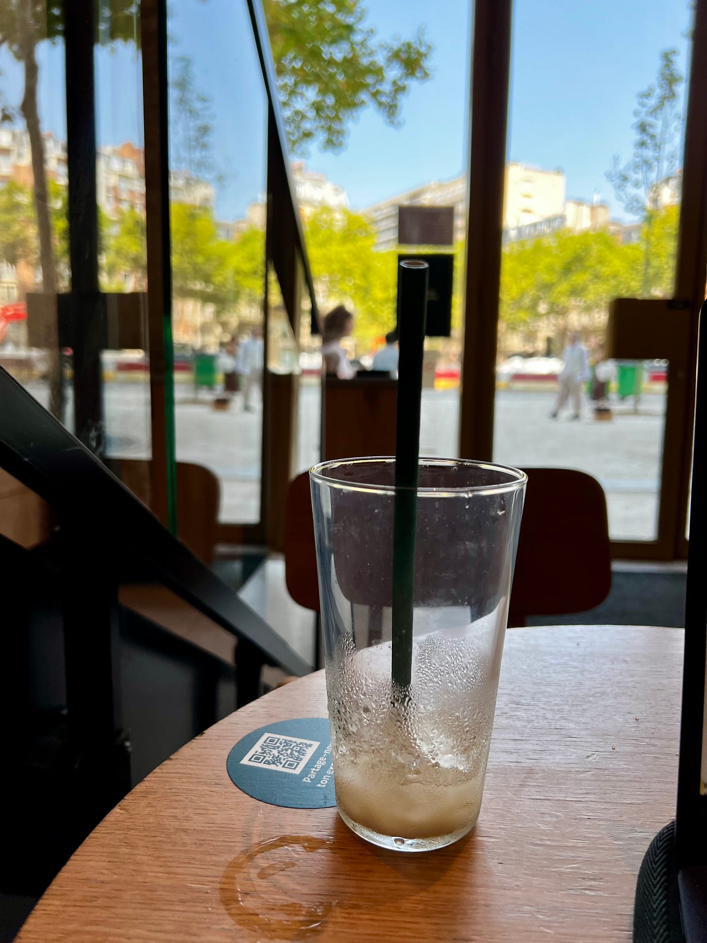 Half-finished iced coffee with straw on wooden table inside café, with blurred street view through windows.