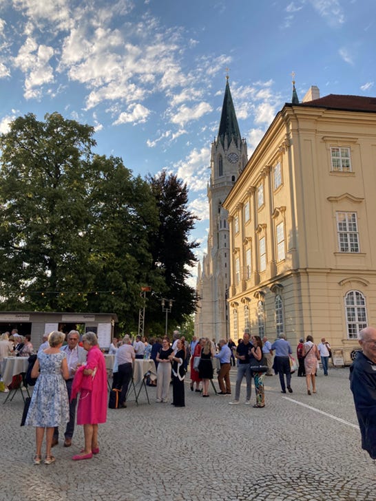 Guests gathering outdoors at Klosterneuburg Abbey beneath a blue sky filled with sunlit clouds, with the abbey tower rising behind them.