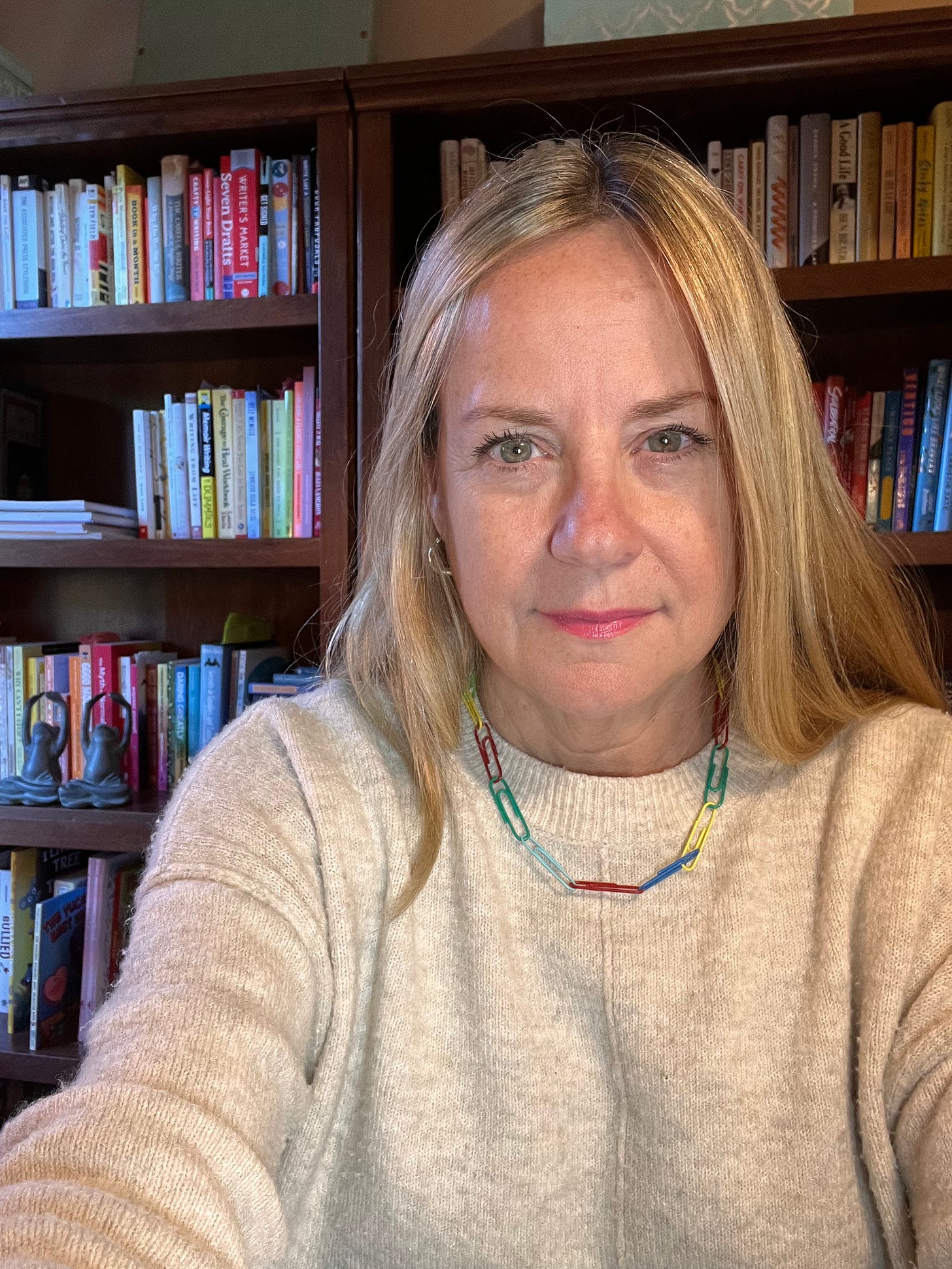 A blonde woman wearing a white sweater and a paperclip neclace sits in front of a bookcase