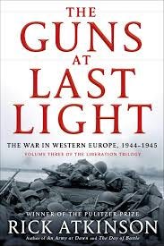 A wartime photo in faded sepia tones shows a group of Allied troops climbing over an obstacle.  The title “The Guns at Last Light” appears in large, capitalized red serif font centered near the top, with Rick Atkinson's name below in smaller white text. The background evokes dusk or early morning light, reinforcing the “last light” motif.