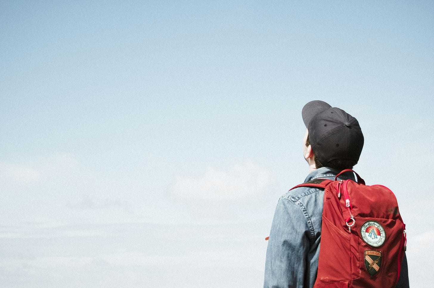 A person wearing a red backpack and baseball cap stands facing an open sky, viewed from behind.