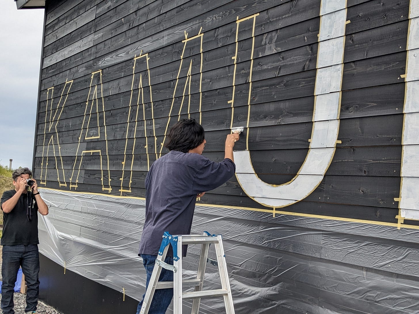 Takamasa from Miraku Ramen painting Kamui name on exterior warehouse wall at Rishiri distillery