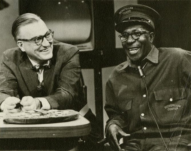 White man in bow tie at desk and black man in Rockefeller Center elevator operator uniform laugh together