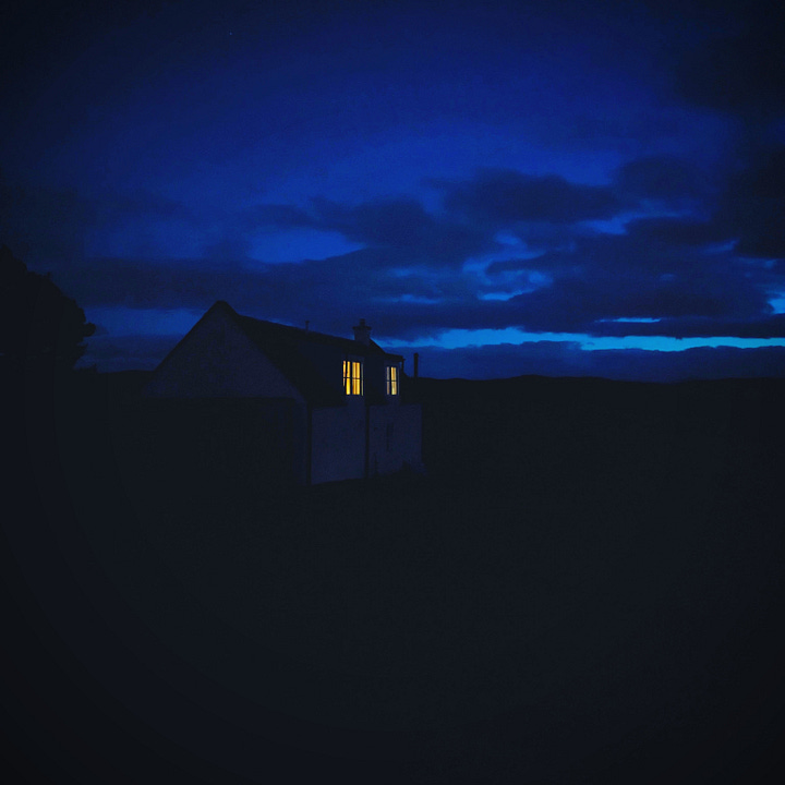 Image 1 is a photo of a cottage in the gloaming, two yellow windows piercing the shadows under the fading blue sky. Image 2 is a photo of a forestry track lined by pine trees, a ray of sunshine bursting through the branches.