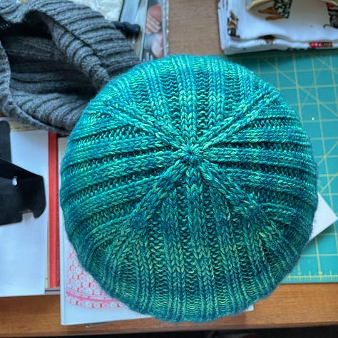 A hand knit hat in teal yarn; an identical hat in grey yarn; a woman with glasses wearing a grey watch cap.