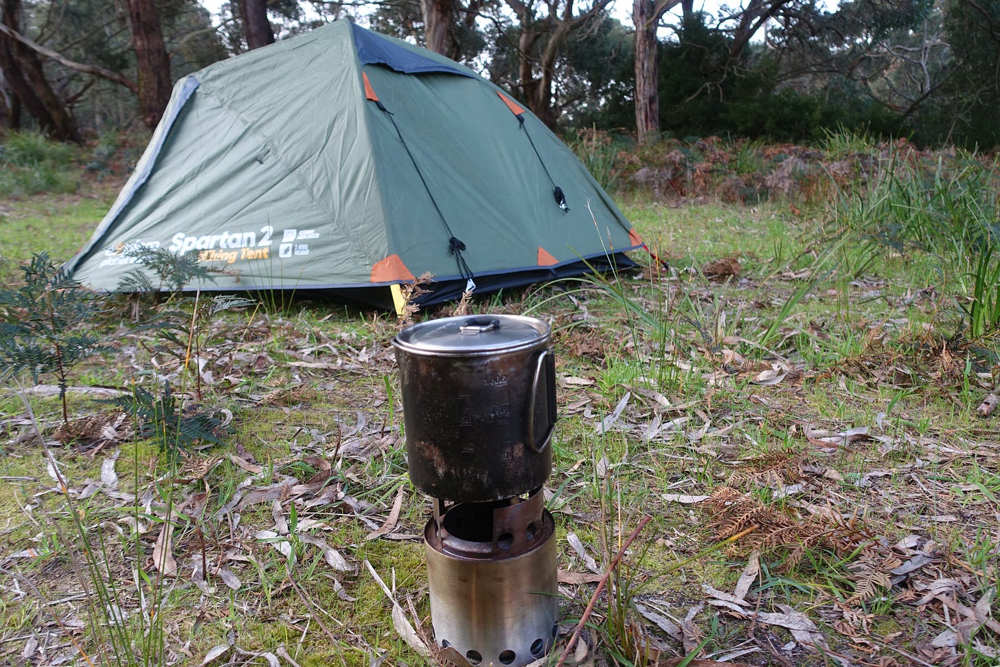 Green Spartan 2 hiking tent set up in eucalyptus forest with camping stove and pot in foreground on leaf-covered ground