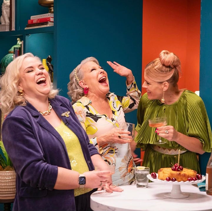 Busy, Caissie and Bette Midler are posing on the set of Busy This Week. Busy is wearing a pleated olive green evening gown. Caissie is wearing a navy velour suit with a lime green shell under the jacket. Bette is wearing a Pucci top in white, blue, black and yellow with white pants. In one photo, they sit smiling. In one photo Busy is posing playfully with her back to the camera. In another, Caissie poses alone to show off her suit. In the final photo, the trio are at the murphy bar on set and they are sharing a huge laugh.