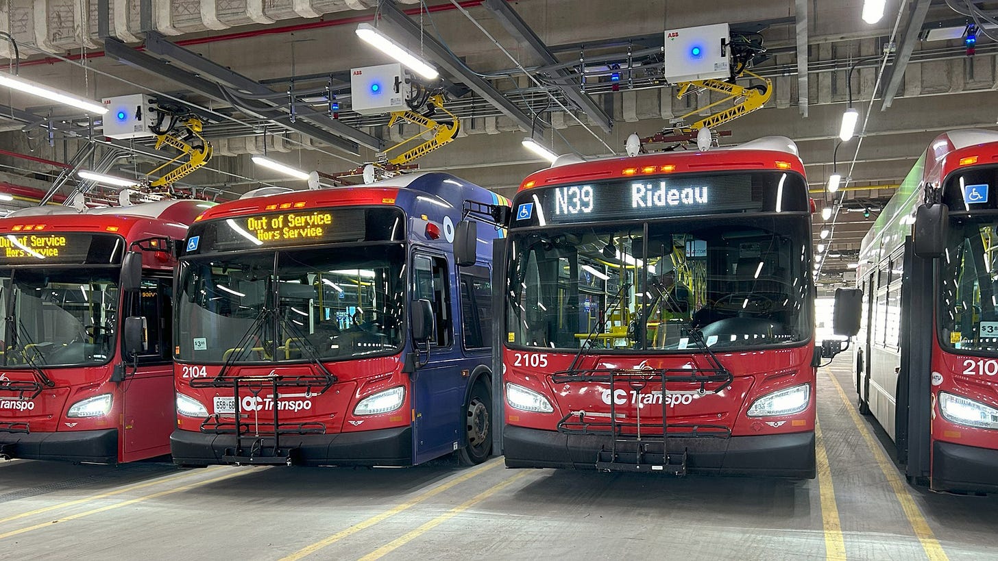 Four buses parked inside an OC Transpo garage. One of the four is a new electric bus
