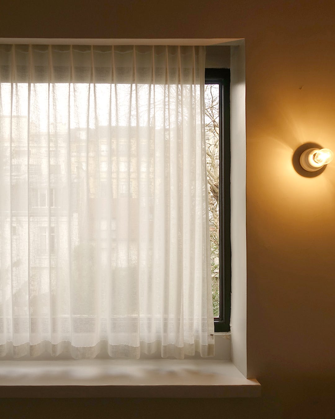 Soft natural light filtering through sheer white curtains at a window alcove, with a glowing wall sconce creating warm ambient lighting in a neutral-toned interior space" Instagram: "Dreamy natural light through sheer curtains with warm wall sconce glow