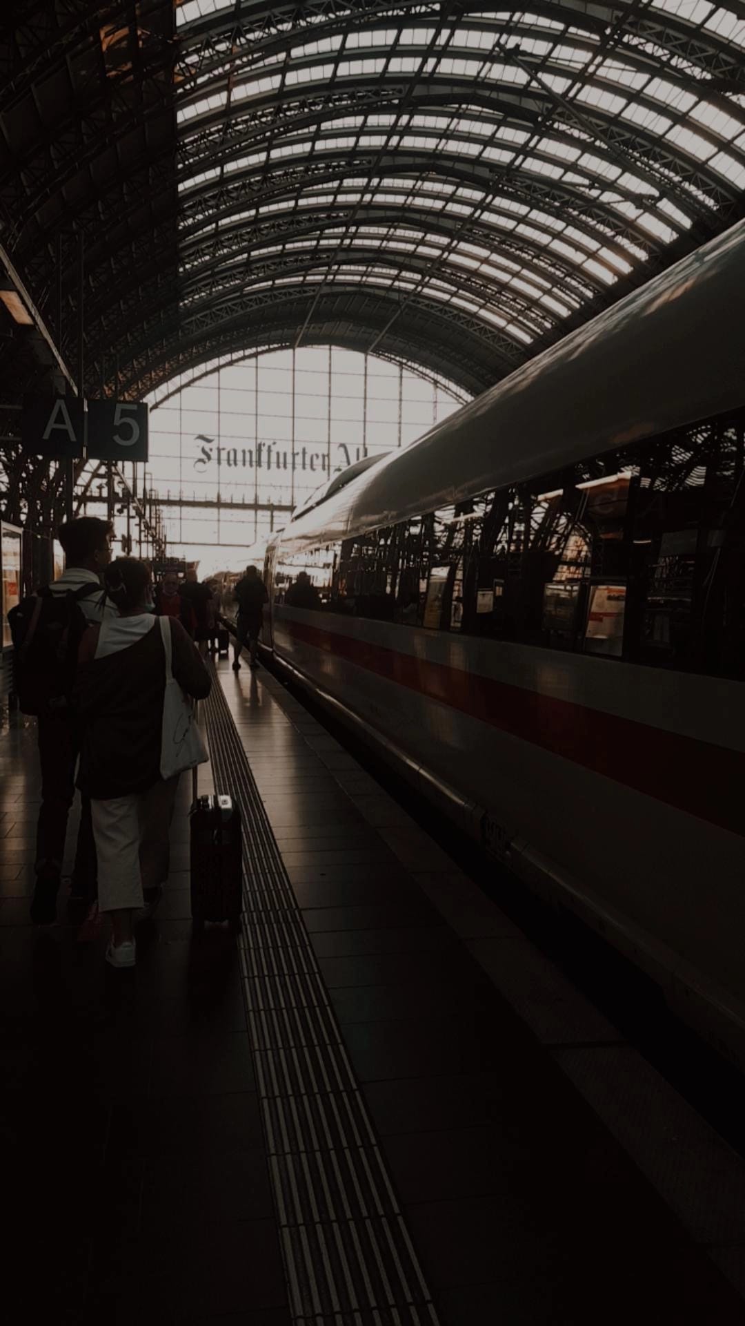 A perspective shot down the platform of a train station in the evening. people are silhouetted and the words 'Frankfurter' are visible on glass windows at the exit of the station